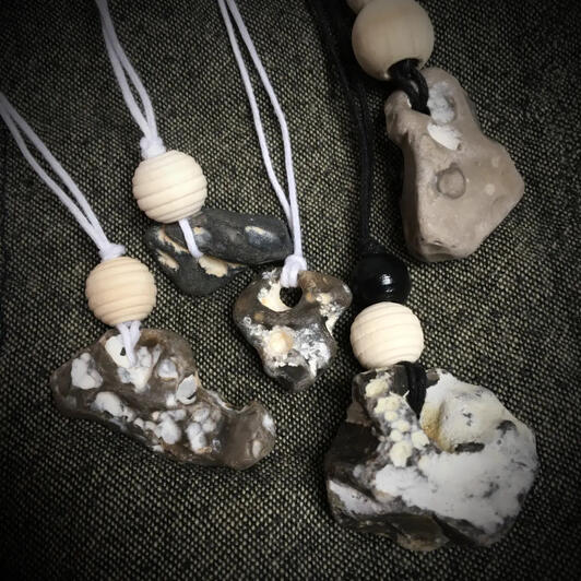 Five natural fairy stones in shades of brown and grey, each with naturally formed holes and organic shapes. Strung on black and white cords to wear as handmade necklaces or lucky charms. Some adorned with natural and black wooden beads. Artisan beach stone