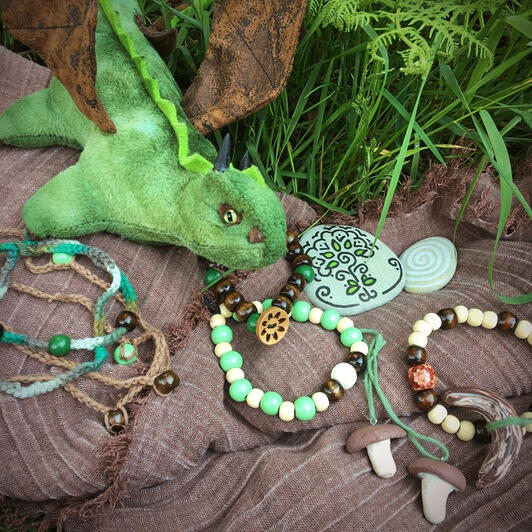 A green dragon plushie with brown wings is placed under a green fern in the grass. Presented on a brown scarf are painted stones with tree symbols, and some jewelry in earthy and green colours. Two pendants shaped like bolete mushrooms, bracelets made of w