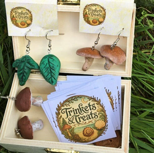 Three pairs of earrings are presented in a wooden box. One is shaped like green leaves, the other two have pendants in the shape of bolete mushrooms. A green dragon plushie with brown wings is placed in the background, sitting in the green grass.