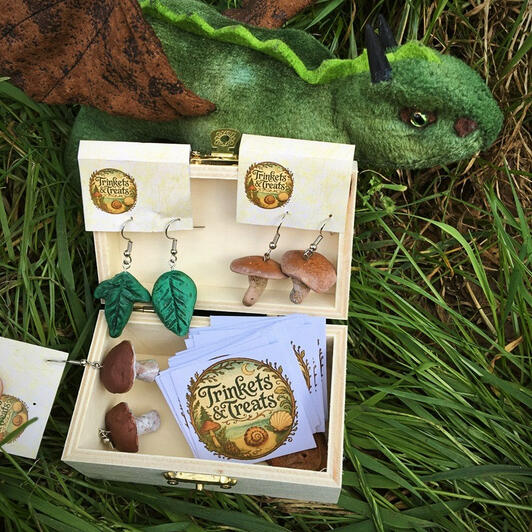 Three pairs of earrings are presented in a wooden box. One is shaped like green leaves, the other two have pendants in the shape of bolete mushrooms. A green dragon plushie with brown wings is placed in the background, sitting in the green grass.