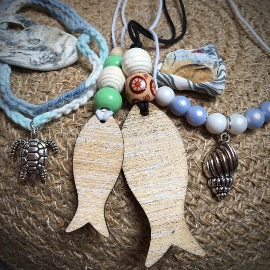 A collection of ocean-inspired jewellery pieces: two wooden fish pendants in light wood with natural and seaweed-green beads, a multicoloured stone pendant in a grey-blue setting, a pale blue and pearlescent bead necklace with a silver snail shell pendant,
